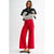 A model stands against a light gray studio background wearing bright red wide-leg cropped pants, a white long-sleeve top with black polka dots, and a black cardigan draped over her shoulders.