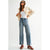 A model wearing a pair of high-waisted, wide-leg light blue denim jeans, styled with a white tank top, an open cream-colored jacket, and brown sneakers, set against a plain light gray studio background.