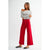 A woman modeling a white off-the-shoulder long-sleeve blouse with black polka dots and bright red wide-leg cropped pants, paired with black flat shoes, against a plain light gray studio background.
