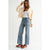 A woman modeling a pair of high-waisted, wide-leg light blue denim jeans, styled with a white tank top, a cream jacket, and red sneakers with white stripes, set against a plain light gray studio background.
