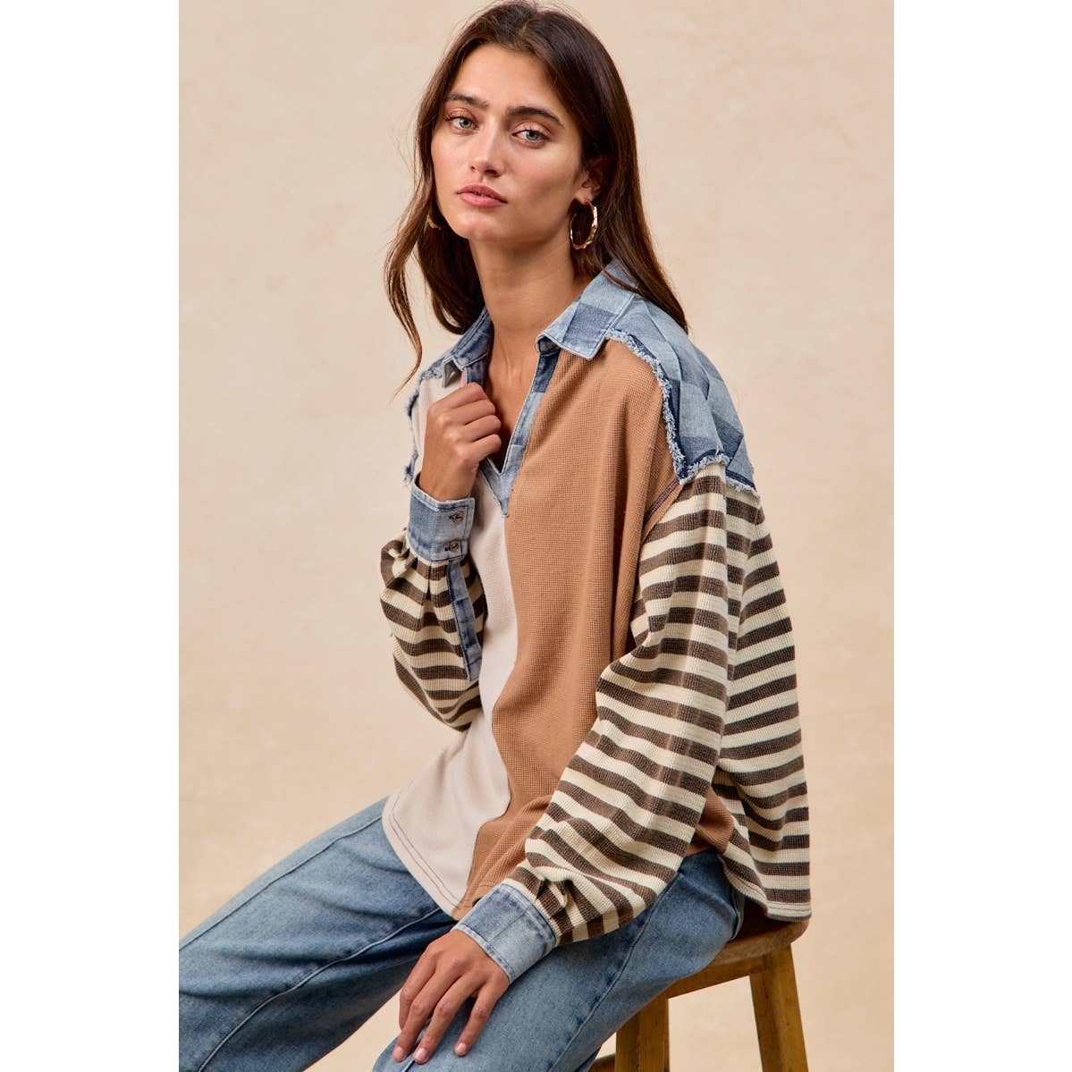 Woman wearing a layered outfit with a denim shirt, beige sweater, and striped sleeves against a beige background.