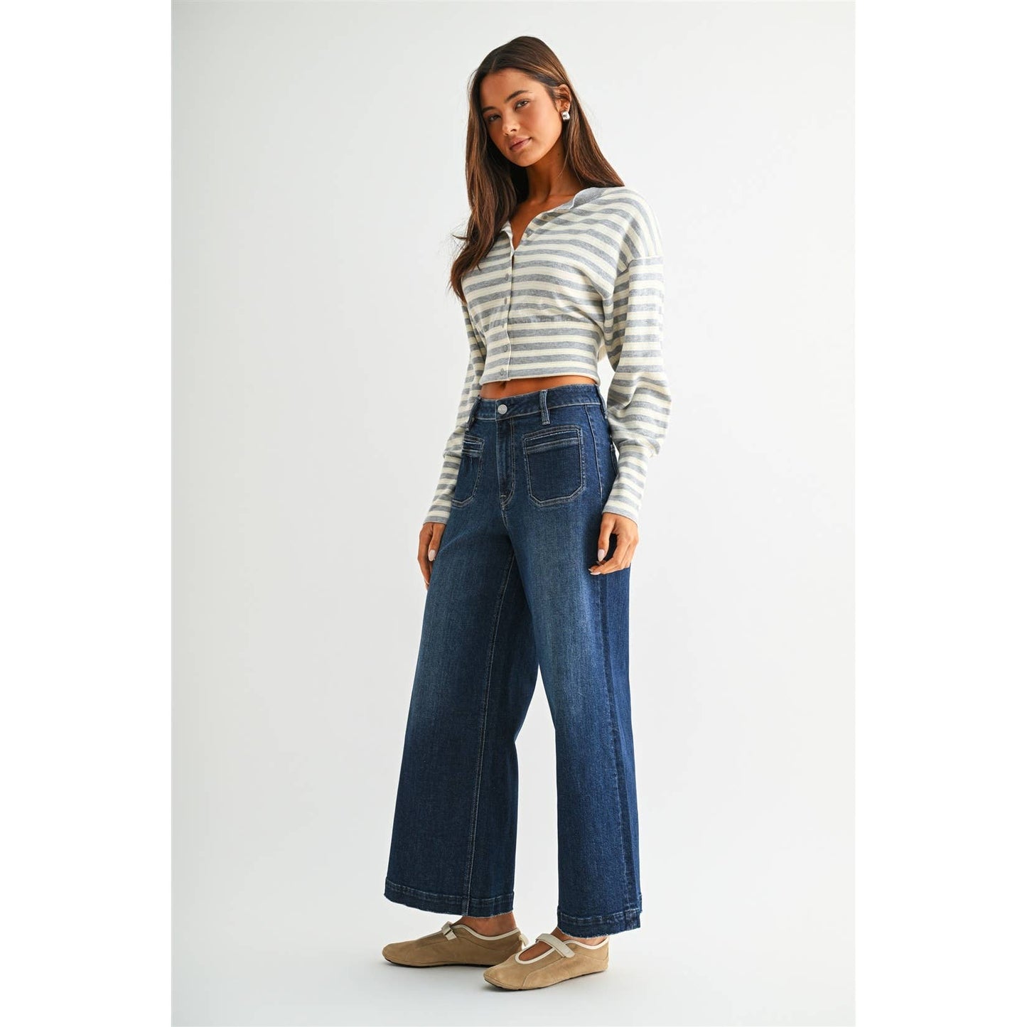 A woman modeling a cropped, long-sleeve striped knit top in gray and white with dark blue wide-leg jeans and beige flats, standing against a light gray studio background.
