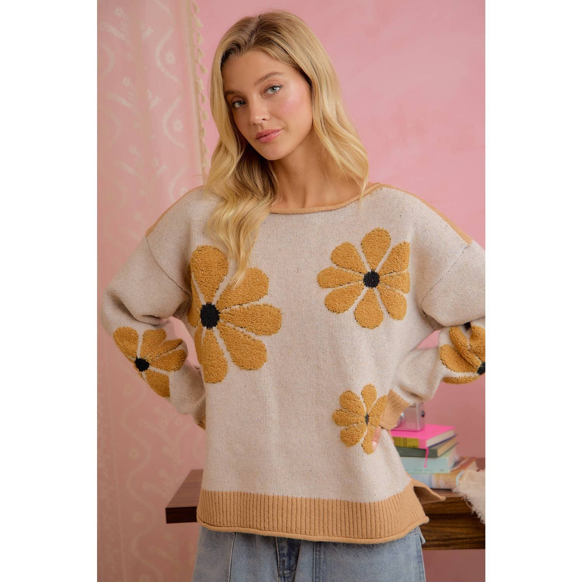 A woman wearing a beige knit sweater with large yellow daisy flower appliqués and ribbed trim, standing against a pink patterned wall.