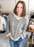 A smiling woman with shoulder-length brown hair stands in a clothing boutique wearing a gray and white patterned knit sweater with peach stripes and blue jeans.