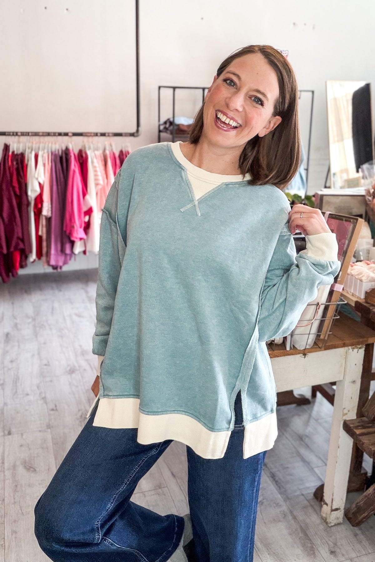 A woman smiling in a clothing boutique, wearing an oversized sage green sweatshirt with cream ribbed trim over a white crewneck tee and blue jeans.