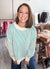 A smiling woman with shoulder-length brown hair wearing a cream and mint green striped knit sweater and blue jeans, standing in a boutique setting.