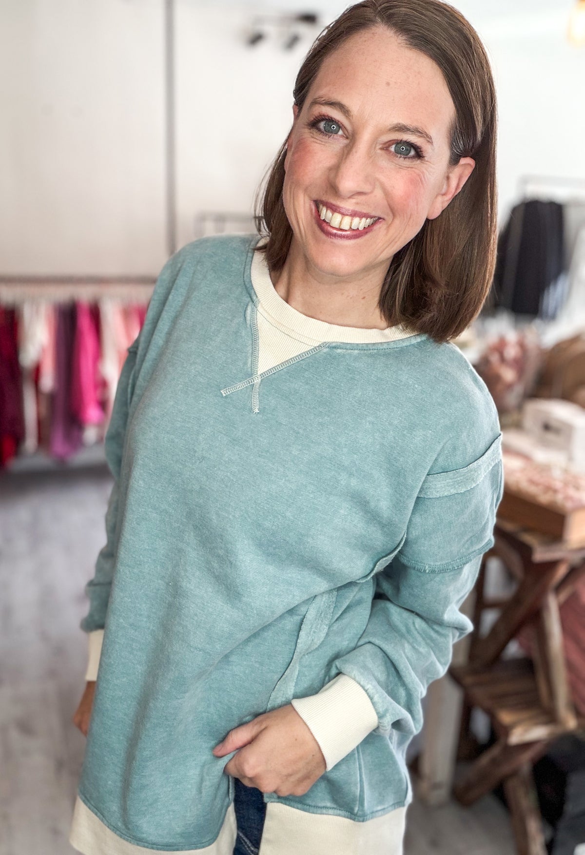 A smiling woman with brown hair is wearing a dusty teal long-sleeve knit top over a cream undershirt, standing in a clothing boutique.