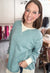A smiling woman with brown hair is wearing a dusty teal long-sleeve knit top over a cream undershirt, standing in a clothing boutique.
