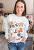 A smiling woman wearing a light gray crewneck sweatshirt with a colorful Halloween-themed graphic print, paired with blue jeans in a retail store setting.