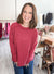 A smiling woman with shoulder-length brown hair wearing a ribbed dusty rose long-sleeve sweater and blue jeans, standing in a boutique setting with clothing racks and wooden tables in the background.