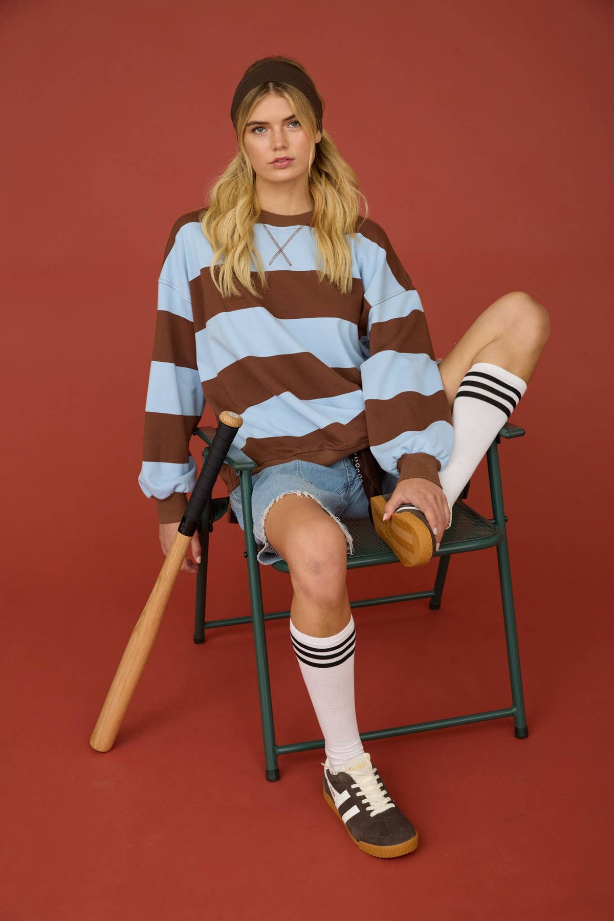A woman sits on a green folding chair against a solid terracotta red studio background, wearing a brown and light blue striped long-sleeve sweatshirt, denim shorts, white knee-high socks with black stripes, and brown sneakers. She holds a wooden baseball bat.