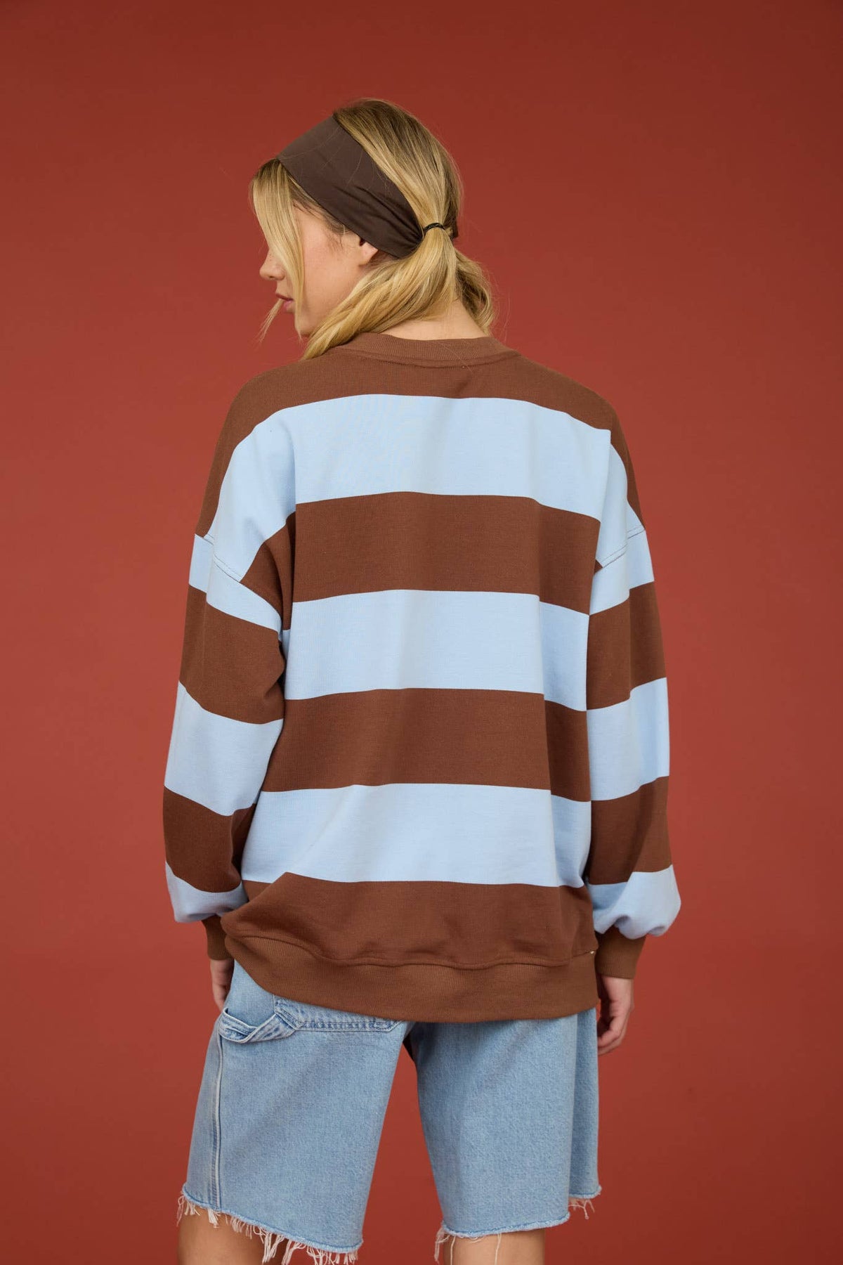 A woman shown from the back wearing an oversized long-sleeve sweatshirt with horizontal brown and light blue stripes, paired with light-wash denim shorts, against a solid terracotta red background.