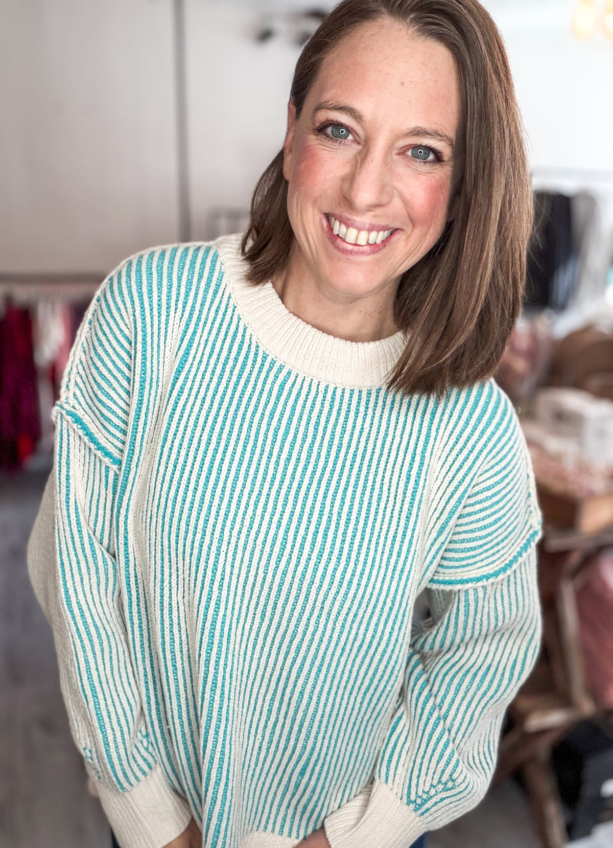 A smiling woman with shoulder-length brown hair wearing a cream and turquoise striped knit sweater, photographed in a softly blurred indoor setting.