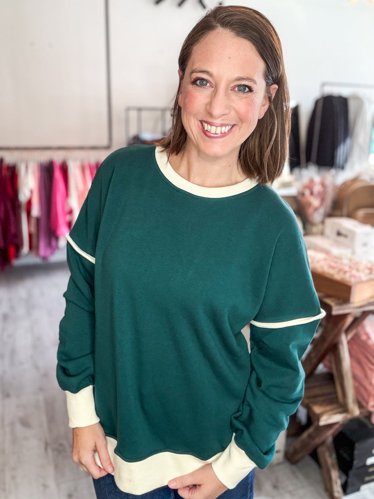 A woman smiling while wearing a dark green long-sleeve crewneck sweatshirt with cream ribbed trim at the collar, cuffs, and hem, standing in a clothing boutique.