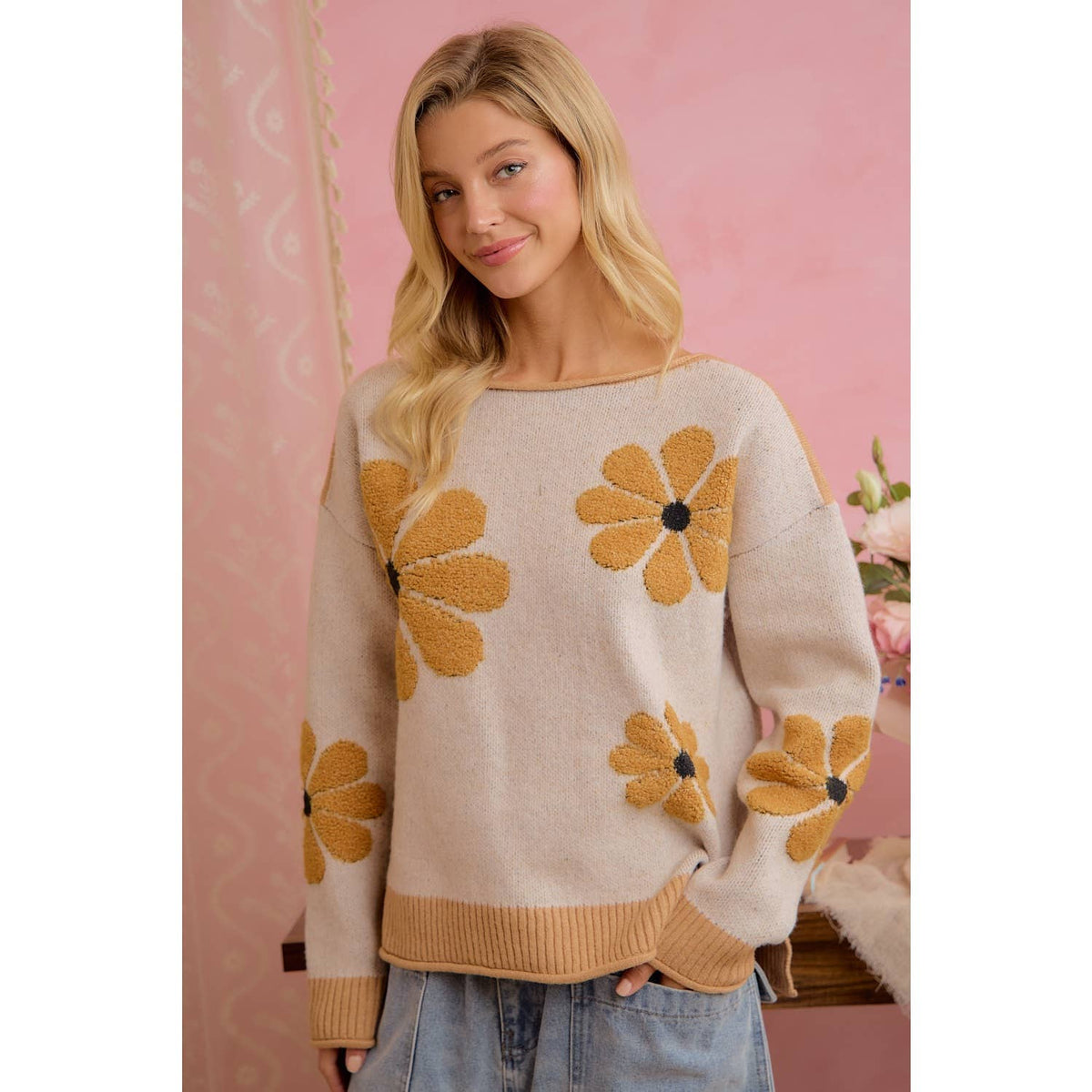 A woman wearing a cream-colored knit sweater with large mustard yellow daisy patterns, paired with light blue jeans, standing against a pink patterned backdrop.