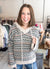 A smiling woman with brown hair stands in a clothing store wearing a gray and cream knit hoodie with a pinkish-beige geometric pattern and blue jeans.