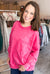 A woman with shoulder-length brown hair smiling in a boutique, wearing a bright pink mineral-wash long-sleeve sweatshirt with a front pocket and blue jeans.