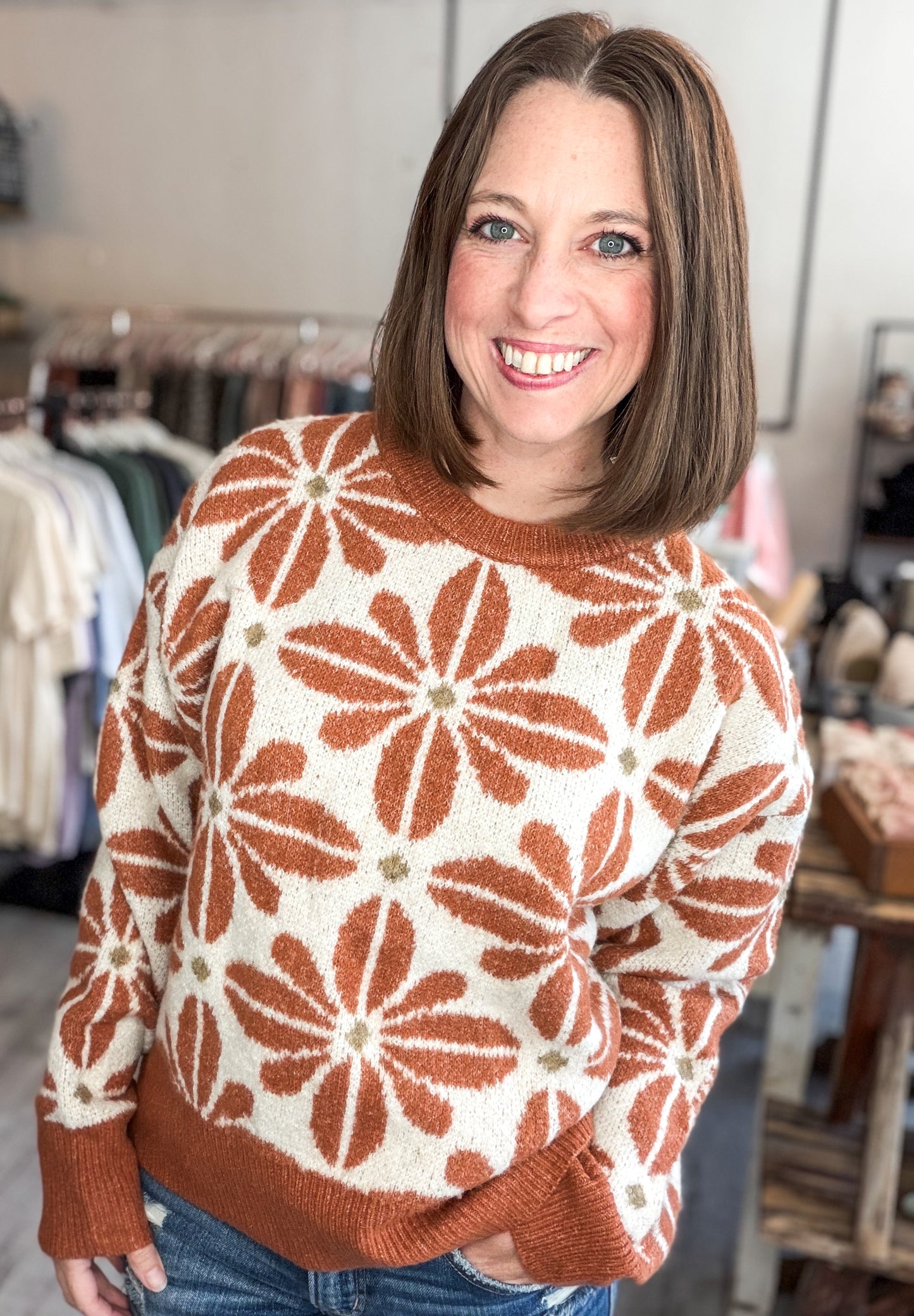 A smiling woman with shoulder-length brown hair wearing a cream knit sweater with a rust-brown floral pattern, standing in a clothing store.