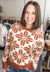A smiling woman with shoulder-length brown hair wearing a cream knit sweater with a rust-brown floral pattern, standing in a clothing store.