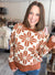 A smiling woman with shoulder-length brown hair models a fuzzy knit crewneck sweater featuring a large rust-orange and cream floral pattern, paired with blue jeans in what appears to be a clothing store.
