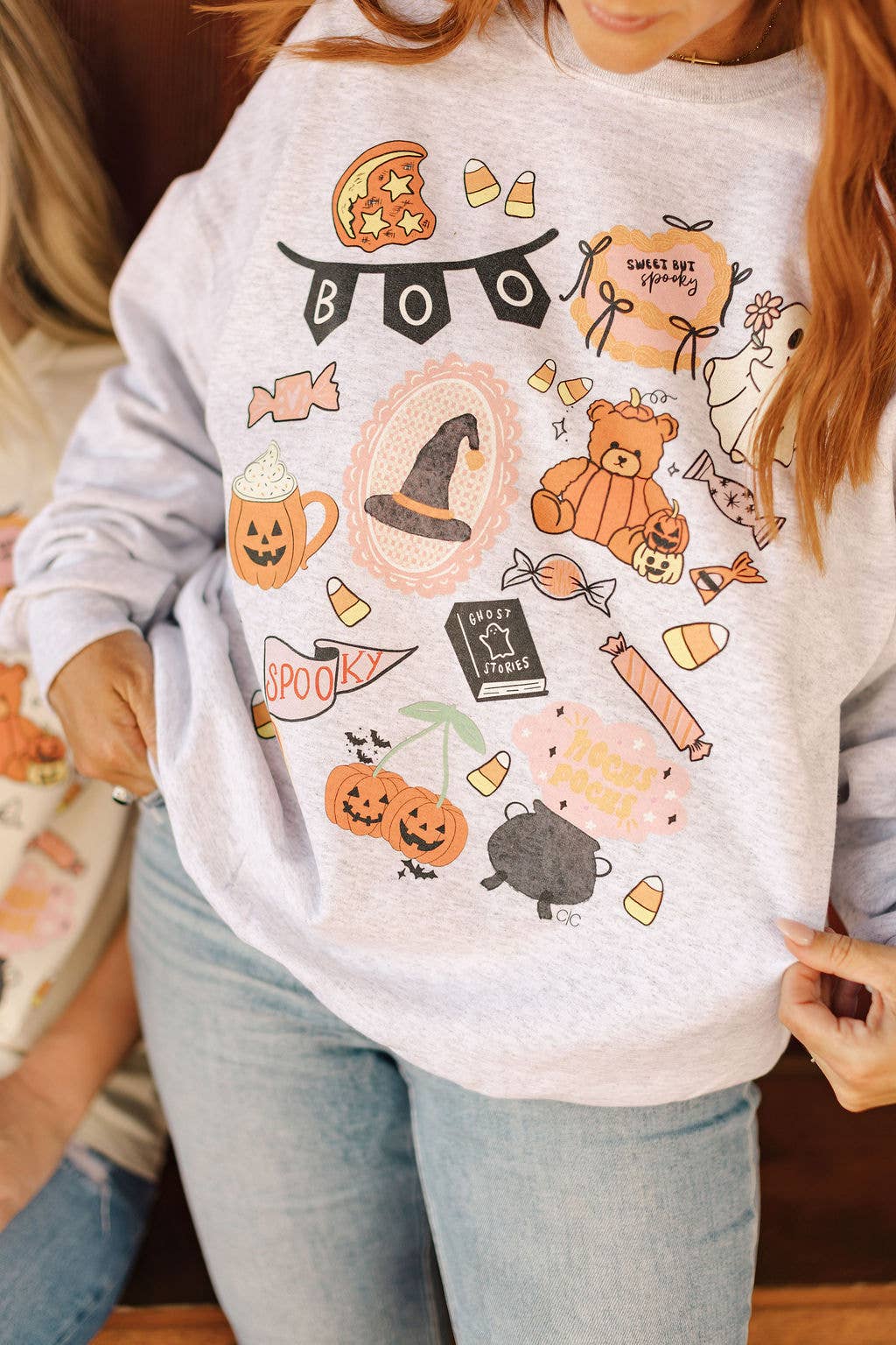 A person wearing a light heather gray sweatshirt with a colorful Halloween-themed graphic print featuring pumpkins, candy corn, a witch hat, a teddy bear, a cat, and the word 'BOO'.