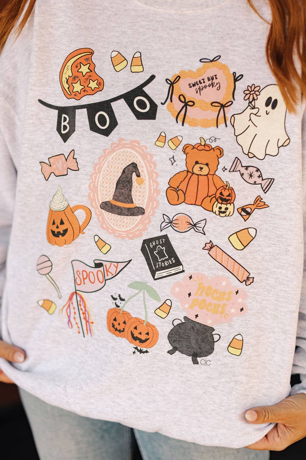 A close-up of a person wearing a light heather gray graphic T-shirt with a colorful Halloween-themed print featuring candy, pumpkins, a witch hat, a ghost, a teddy bear, and the words 'BOO' and 'Sweetest Spooky'.