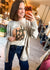 A woman taking a mirror selfie in a boutique, wearing a light gray knit cardigan over a cream graphic t-shirt and dark blue jeans. She holds an orange smartphone in front of her face.