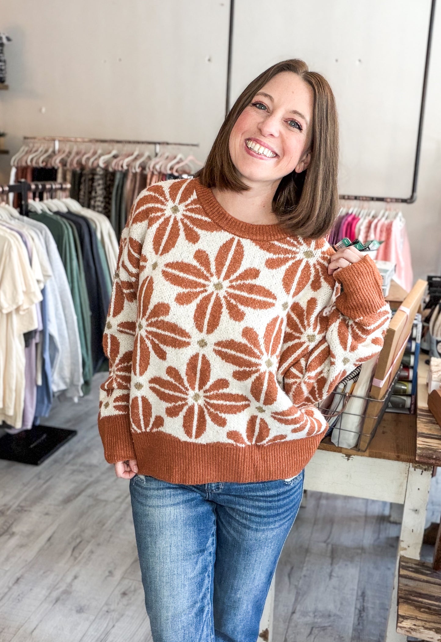 A smiling woman wearing a rust-orange and cream knit sweater with a large floral pattern, paired with blue jeans, standing inside a clothing boutique.