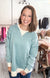 A smiling woman with shoulder-length brown hair stands in a boutique wearing a light blue-green long-sleeve crewneck sweatshirt with cream ribbed trim over a cream top and blue jeans.