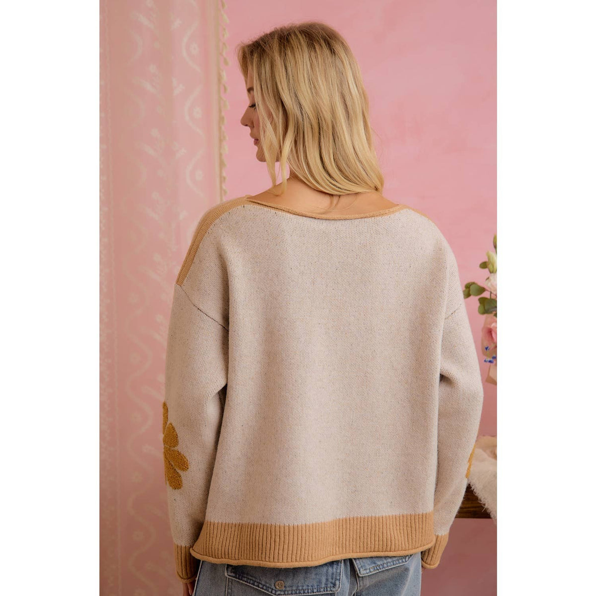 A woman viewed from behind wearing a beige knit sweater with tan ribbed trim and a yellow embroidered flower detail on the sleeve, set against a pink patterned wall.