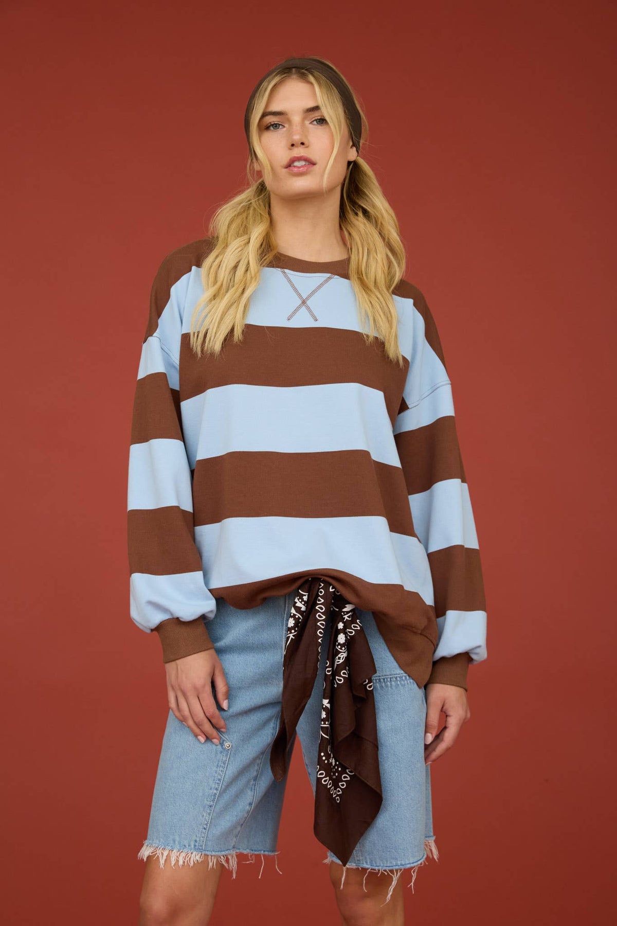 A woman modeling a brown and light blue horizontally striped long-sleeve top with lace-up detail at the chest, paired with denim shorts and a brown patterned bandana, set against a solid terracotta background.