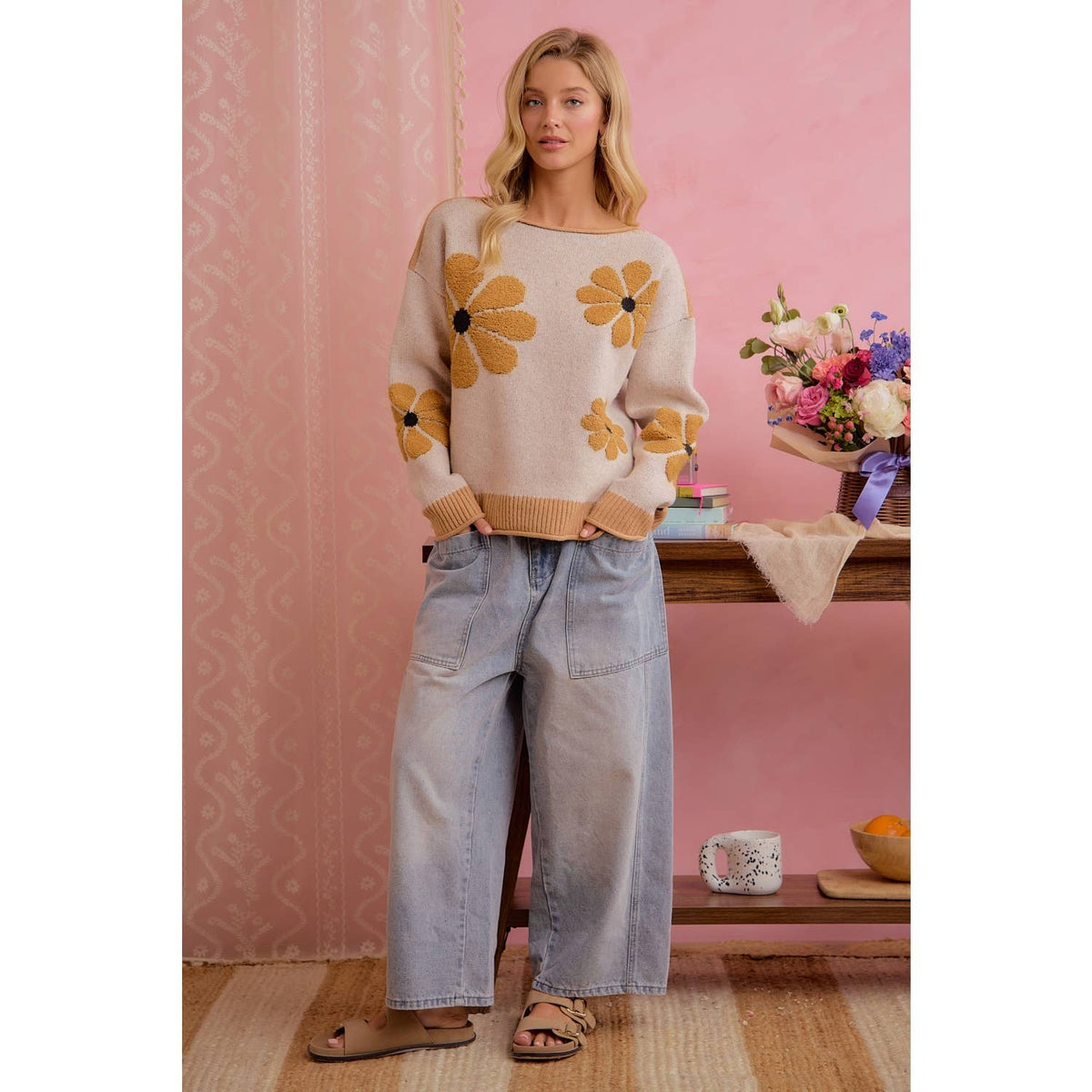 A woman stands indoors wearing a cream knit sweater with large mustard-yellow flower motifs, light-wash wide-leg jeans, and tan sandals. The background is a pink wall with a shelf holding flowers and small decor items.