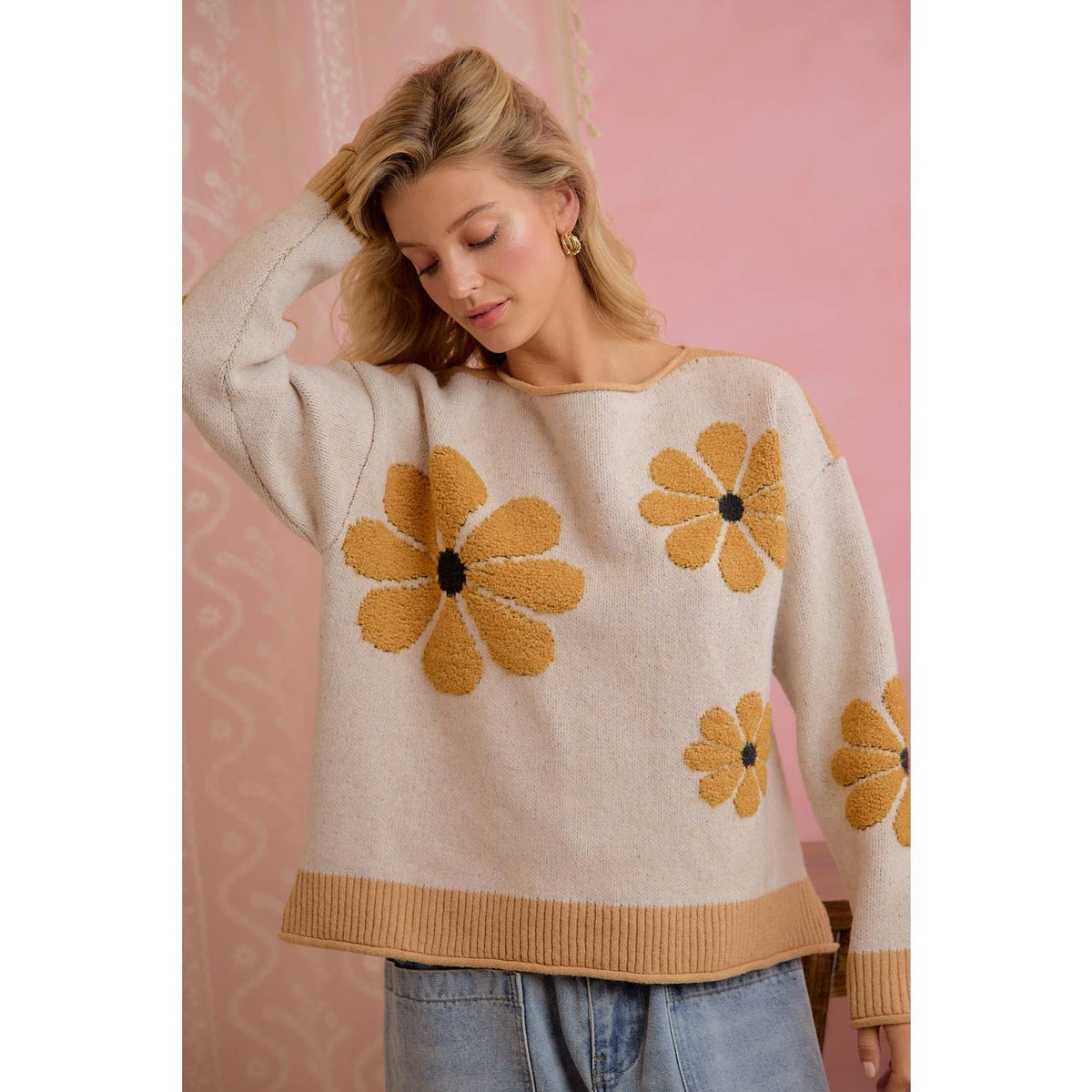 A woman wearing a cream-colored knit sweater with large mustard yellow daisy patterns and a ribbed mustard yellow hem, paired with light blue jeans, set against a pink patterned backdrop.