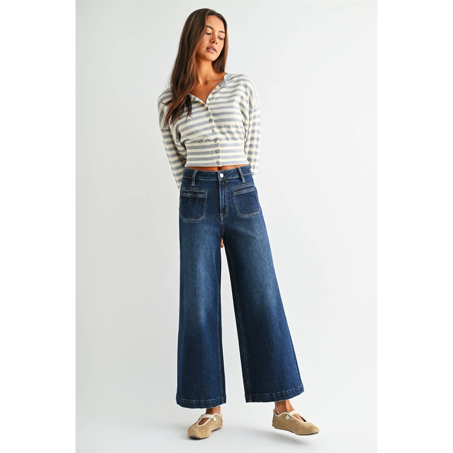A woman modeling a striped long-sleeve crop top and high-waisted, wide-leg blue denim jeans against a plain light gray studio background.