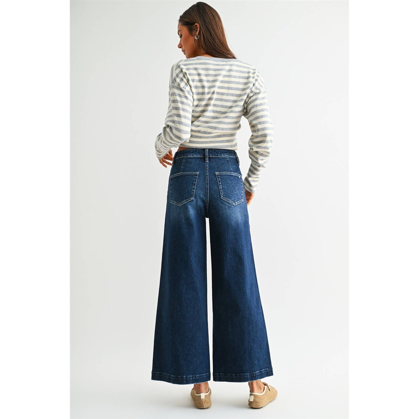 A woman shown from the back wearing a gray and white striped long-sleeve crop top and dark blue wide-leg denim jeans, paired with beige flat shoes, against a light gray studio background.